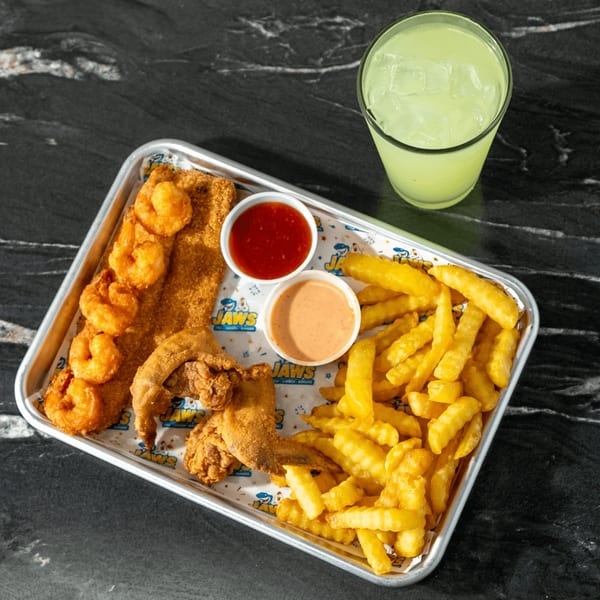 Jaws seafood platter with fried shrimp, fish, chicken, crinkle-cut fries, dipping sauces, and a lime drink on a black table.