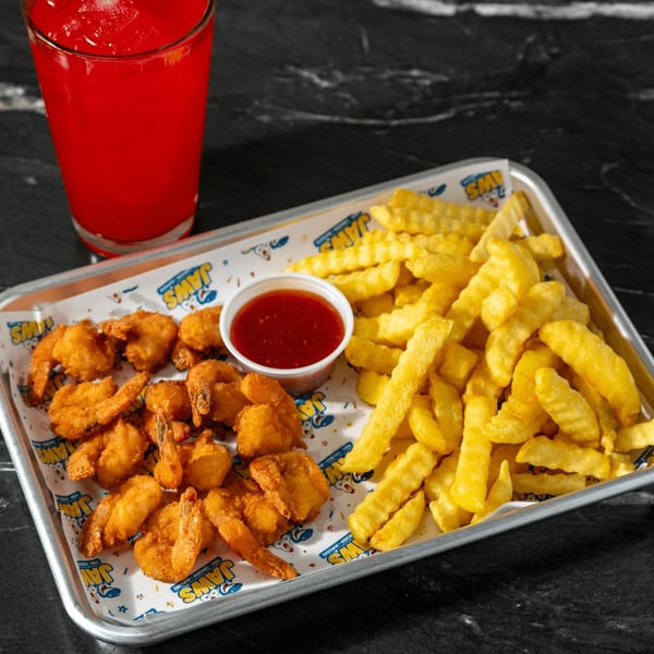 Tray of crispy fried shrimp, crinkle-cut fries, and red dipping sauce beside a red drink on a dark table.