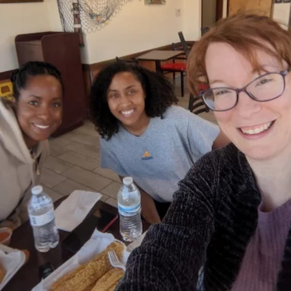 Three smiling women pose for a selfie at Jaws Hot Chicken & Seafood, with fried fish on the table.