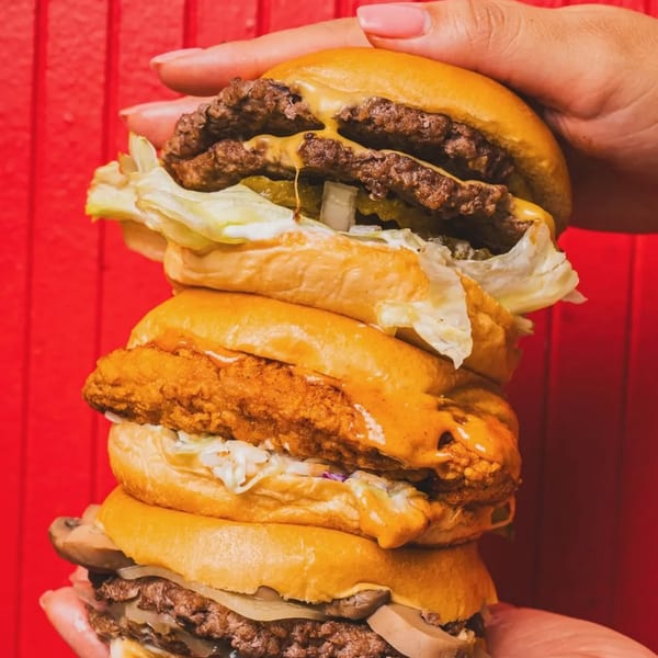 A hand holds a towering stack of three Jaws Hot Chicken & Seafood burgers against a red background.