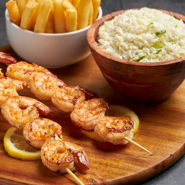 Grilled shrimp skewers with lemon slices, a bowl of fries, and coleslaw on a wooden board.