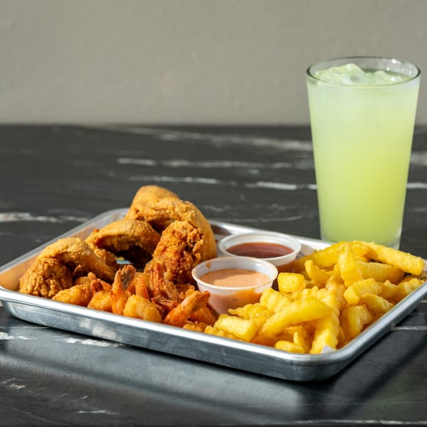 Metal tray of fried chicken, shrimp, fries, dipping sauces, and a tall lime-green iced drink on a dark table.