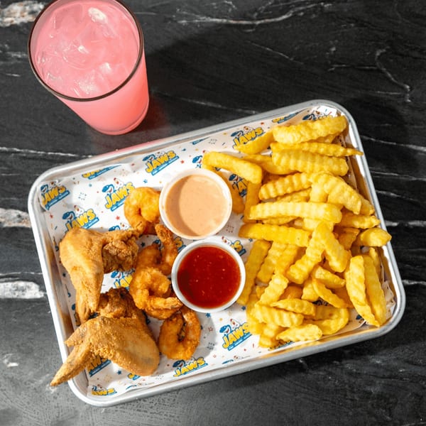 Tray of fried chicken, shrimp, crinkle-cut fries, dipping sauces, and a pink iced drink on a black marble table.