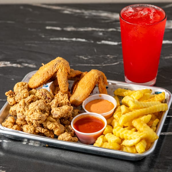 Tray of fried chicken pieces, battered wings, fries, dipping sauces, and a red iced drink on a dark tabletop.