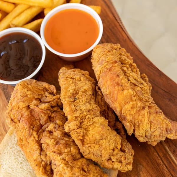 Crispy fried chicken tenders, fries, and two dipping sauces on a wooden board at Jaws Hot Chicken & Seafood.